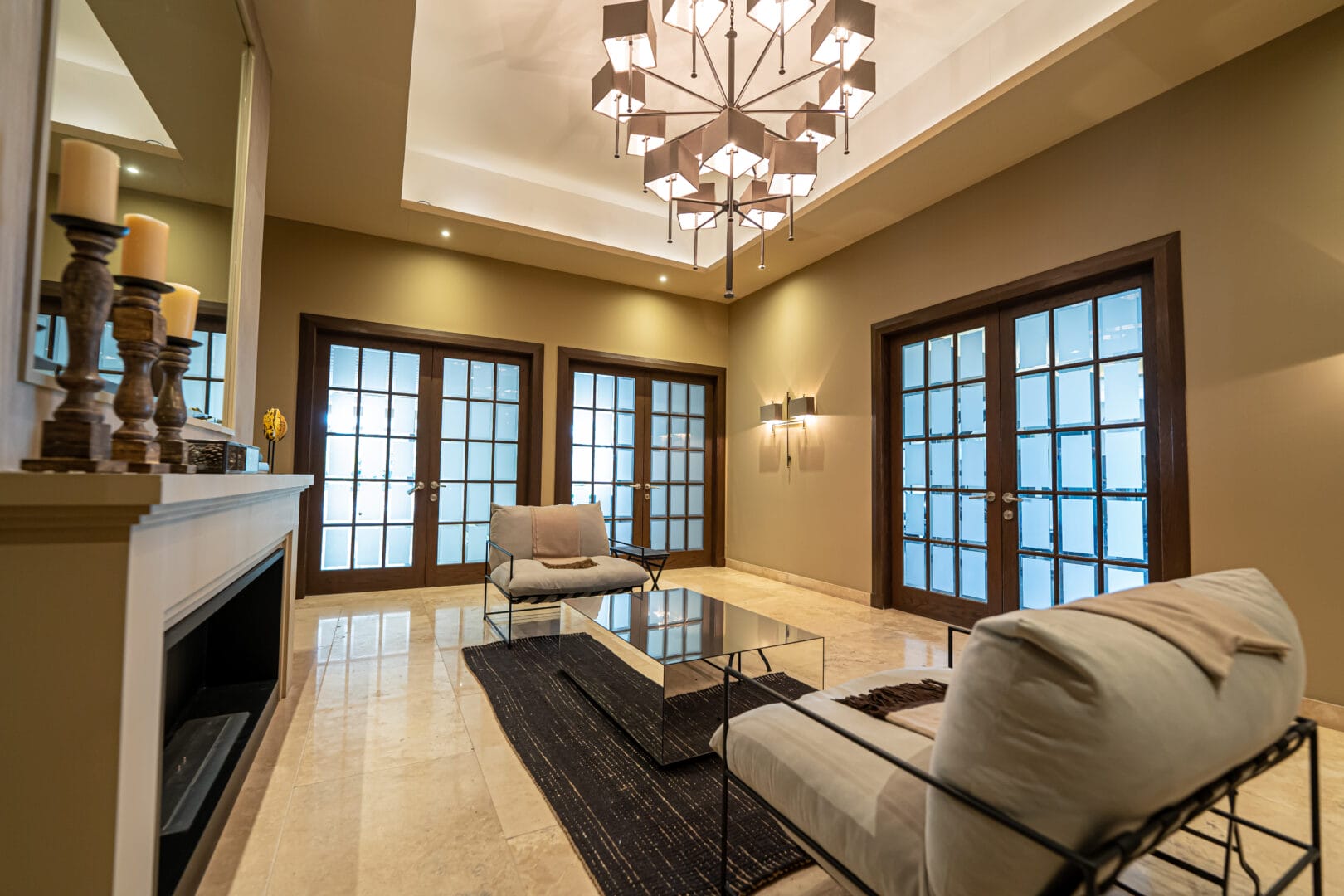 Sala moderna con paredes en tonos beige, dos sillones con estructura metálica y cojines claros, mesa de centro con superficie reflectante sobre alfombra oscura, piso de mármol claro, lámpara de techo con diseño geométrico y tres puertas dobles con marco de madera y vidrio esmerilado al fondo.