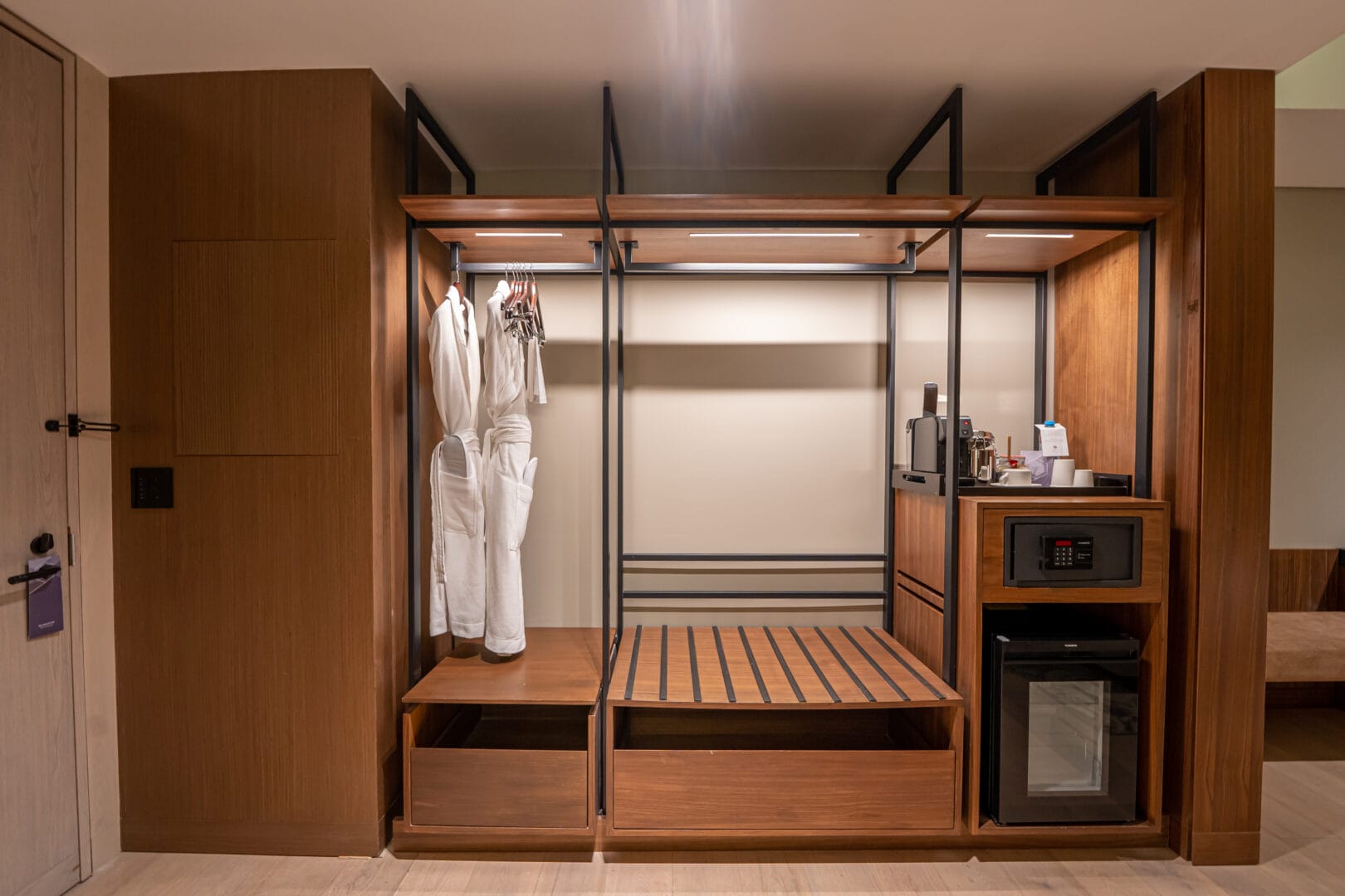 Clóset abierto de madera y metal con dos batas blancas colgadas, espacio vacío con base de madera para equipaje, caja fuerte, mini refrigerador y cafetera con tazas y proveeduría sobre un mueble a la derecha.