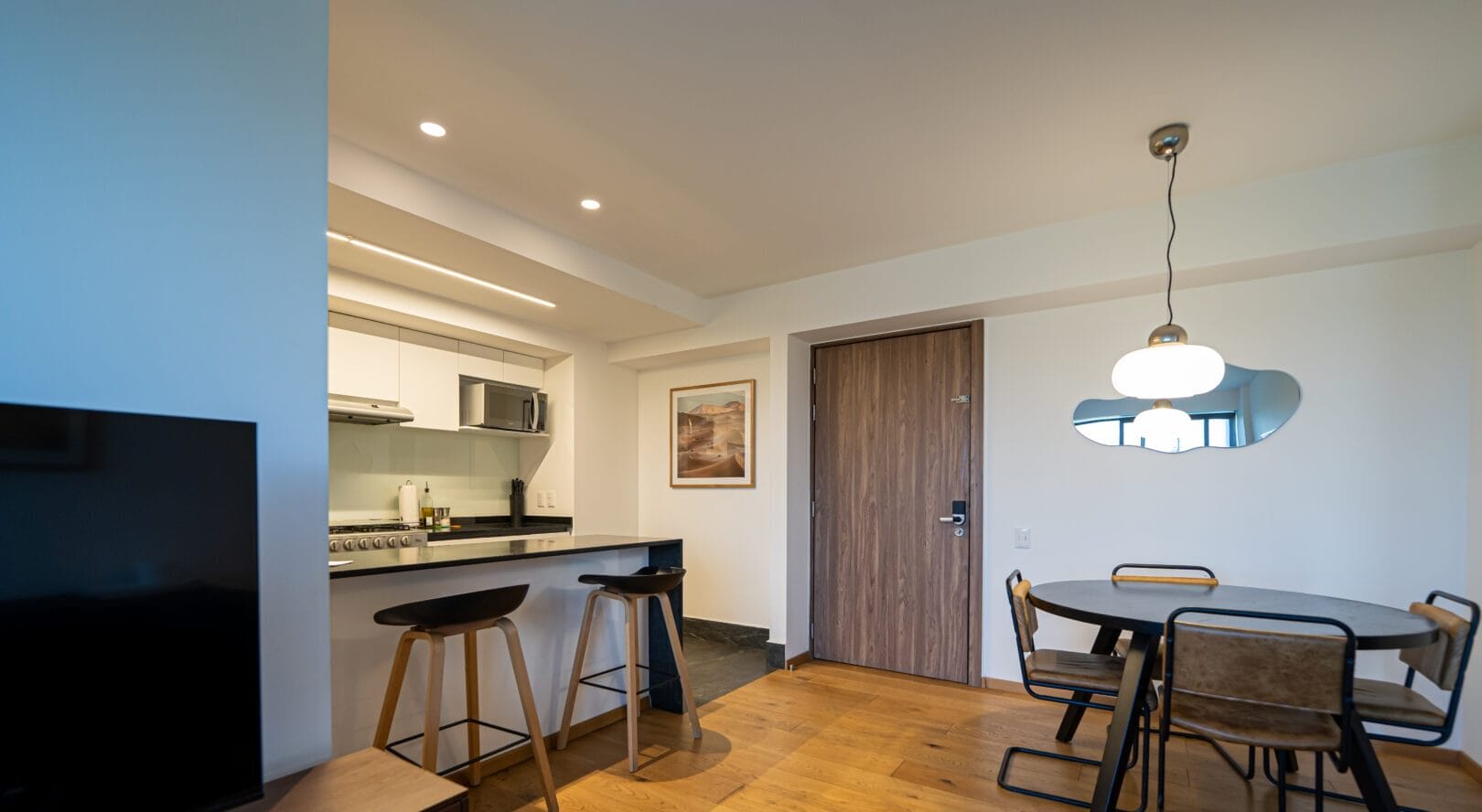 Sala comedor con piso de madera clara, mesa redonda con cuatro sillas de estructura metálica y madera, lámpara colgante blanca y espejo decorativo en pared blanca, puerta de madera, barra desayunadora con dos bancos altos, cocina con gabinetes blancos y microondas empotrado.