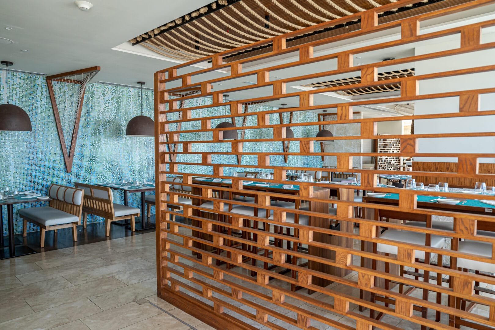 Interiores de restaurante moderno con muro divisor de madera con diseño geométrico, mesas con manteles azules, sillas y bancos tapizados en tonos grises, lámparas colgantes de estilo industrial y pared decorada con mosaicos en tonos azules y verdes.