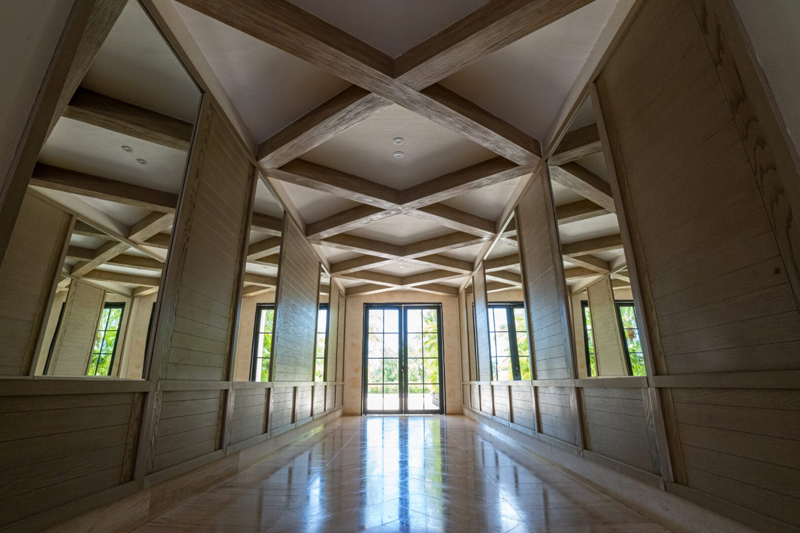 Pasillo amplio con paredes de madera clara y espejos grandes a ambos lados, techo con vigas decorativas en patrón de cruz, y puerta de vidrio al fondo que muestra un jardín exterior con vegetación.