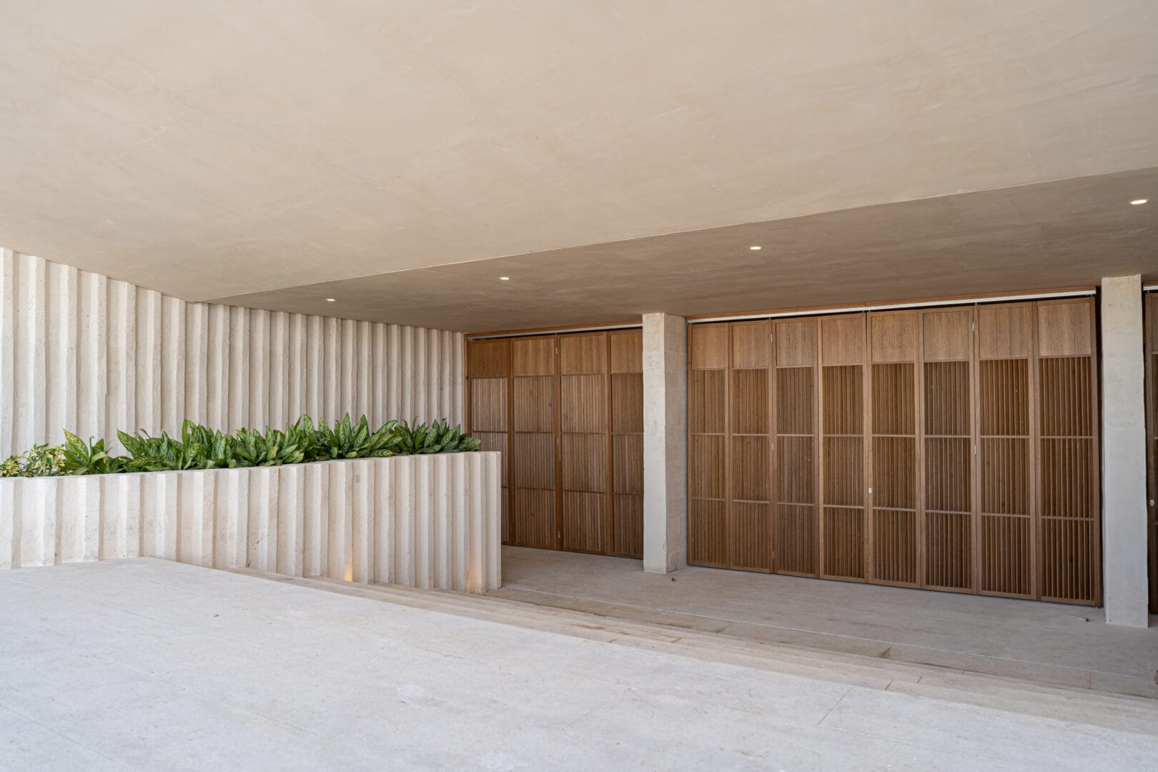 Área exterior moderna con paredes y techo en tonos beige claro, destacando una jardinera con plantas verdes y puertas de madera con diseño de paneles verticales.