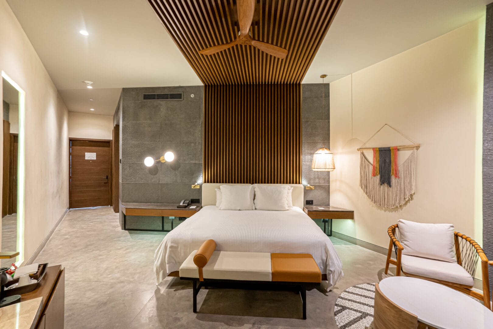 Habitación moderna con cama grande de ropa blanca, dos mesas de noche flotantes con lámparas, muros de tonos gris y beige, techo con paneles de madera y ventilador, banco al pie de la cama y silla con cojín cerca de una mesa ovalada baja.