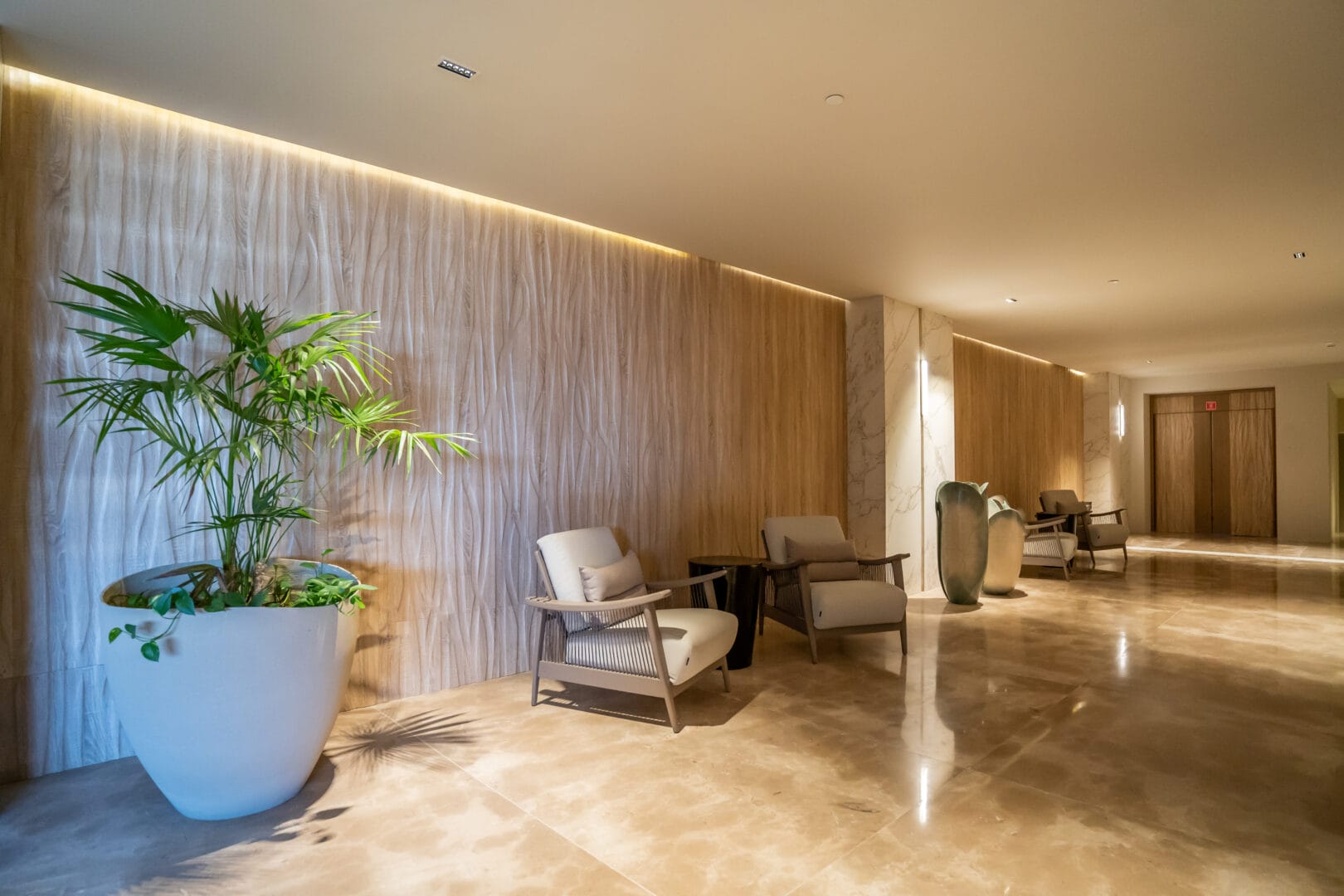 Pasillo moderno y elegante con paredes de madera y mármol, piso de mármol pulido, varias sillas tapizadas beige con estructura de madera, una mesa pequeña y una planta grande en maceta blanca al lado izquierdo.