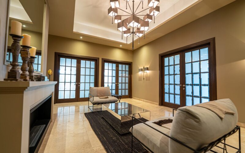 Sala moderna con paredes en tonos beige, dos sillones con estructura metálica y cojines claros, mesa de centro con superficie reflectante sobre alfombra oscura, piso de mármol claro, lámpara de techo con diseño geométrico y tres puertas dobles con marco de madera y vidrio esmerilado al fondo.