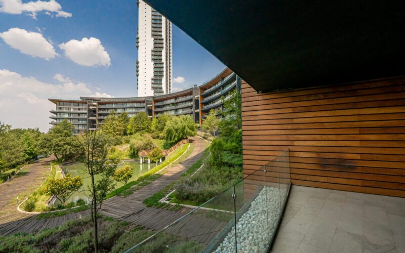 Vista desde un balcón con barandales de vidrio y paredes de madera hacia un jardín con camino de madera, árboles, un pequeño estanque y edificios residenciales modernos de varios pisos bajo un cielo azul con nubes blancas.