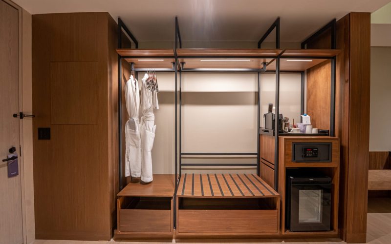 Clóset abierto de madera y metal con dos batas blancas colgadas, espacio vacío con base de madera para equipaje, caja fuerte, mini refrigerador y cafetera con tazas y proveeduría sobre un mueble a la derecha.
