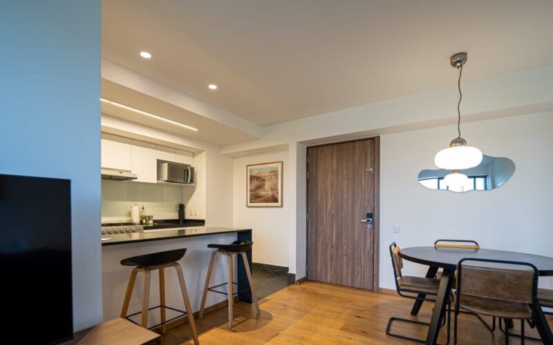 Sala comedor con piso de madera clara, mesa redonda con cuatro sillas de estructura metálica y madera, lámpara colgante blanca y espejo decorativo en pared blanca, puerta de madera, barra desayunadora con dos bancos altos, cocina con gabinetes blancos y microondas empotrado.