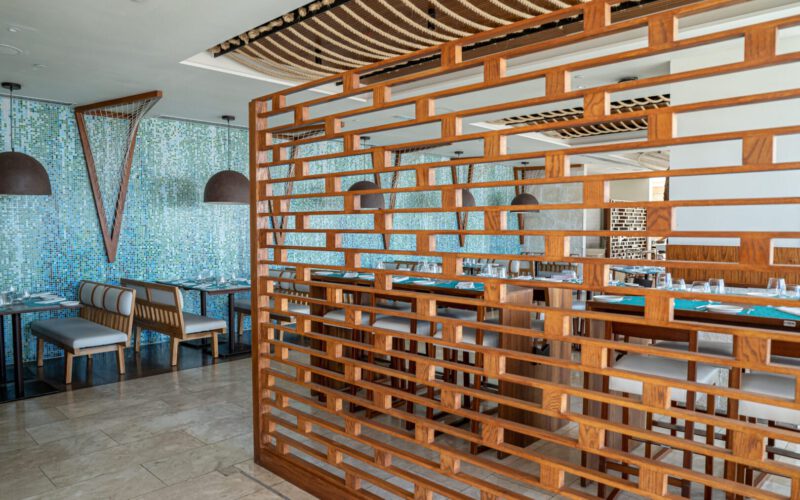 Interiores de restaurante moderno con muro divisor de madera con diseño geométrico, mesas con manteles azules, sillas y bancos tapizados en tonos grises, lámparas colgantes de estilo industrial y pared decorada con mosaicos en tonos azules y verdes.