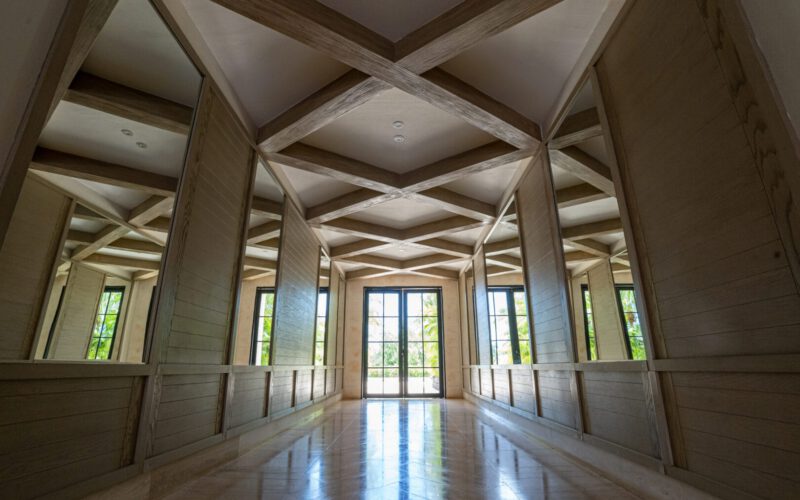 Pasillo amplio con paredes de madera clara y espejos grandes a ambos lados, techo con vigas decorativas en patrón de cruz, y puerta de vidrio al fondo que muestra un jardín exterior con vegetación.