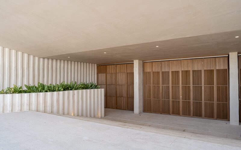 Área exterior moderna con paredes y techo en tonos beige claro, destacando una jardinera con plantas verdes y puertas de madera con diseño de paneles verticales.
