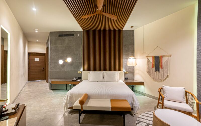 Habitación moderna con cama grande de ropa blanca, dos mesas de noche flotantes con lámparas, muros de tonos gris y beige, techo con paneles de madera y ventilador, banco al pie de la cama y silla con cojín cerca de una mesa ovalada baja.