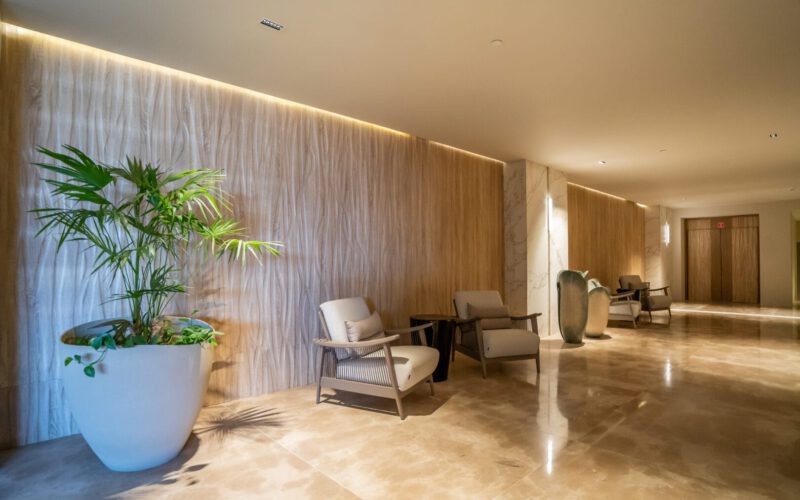 Pasillo moderno y elegante con paredes de madera y mármol, piso de mármol pulido, varias sillas tapizadas beige con estructura de madera, una mesa pequeña y una planta grande en maceta blanca al lado izquierdo.