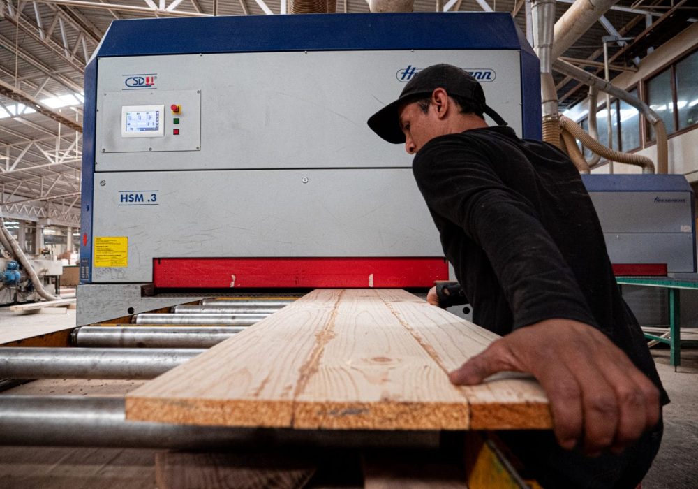 Trabajador con gorra negra manipulando una tabla de madera que entra a una máquina industrial en una carpintería.