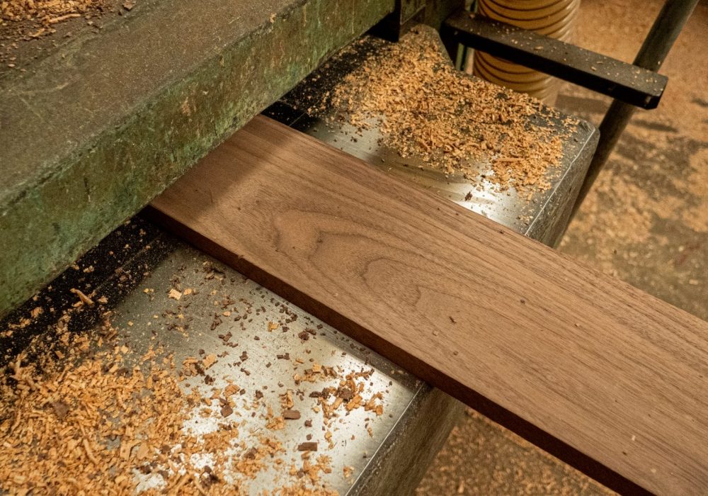 Tabla de madera siendo procesada con máquina cepilladora, rodeada de virutas de madera sobre una superficie metálica y suelo polvoso.