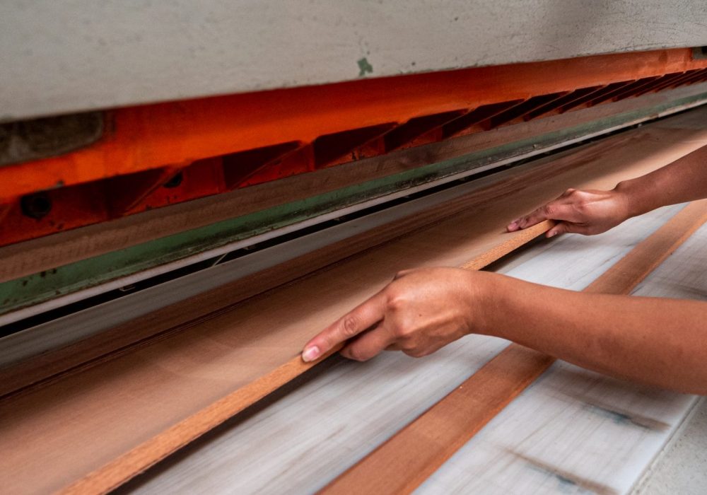 Manos manipulando una tabla de madera en una máquina industrial con partes visibles en color naranja y verde.