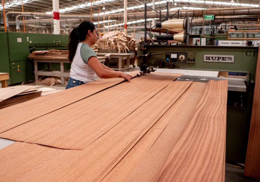 Mujer trabajando en una fábrica con máquina Kuper, manipulando láminas delgadas de madera en una mesa grande dentro de un taller industrial.