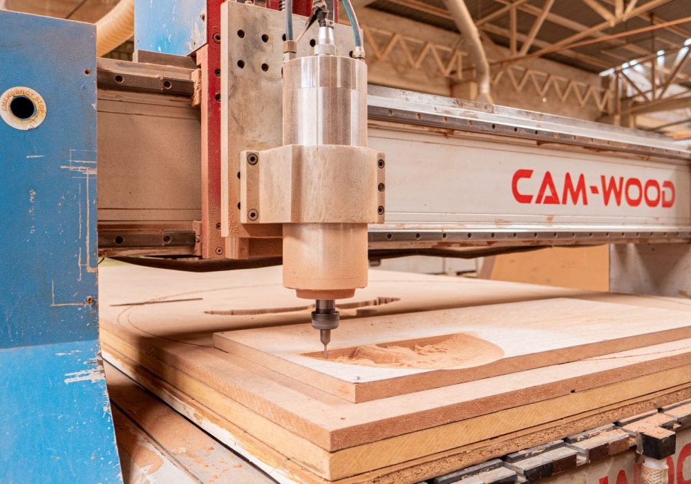 Máquina CNC CAM-WOOD trabajando en corte y tallado de madera en un taller con estructura metálica y vigas en el techo.