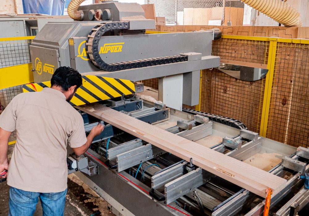 Hombre con camiseta beige y jeans trabajando en una máquina industrial gris y amarilla que procesa madera en un taller.